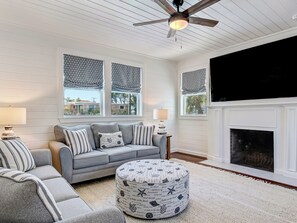 A living room with a grey sofa, patterned ottoman, and a fireplace. - Large Tybee Home for 28 Guests, Private Pool (Tybee Island)