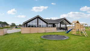 A modern house with large windows, a wooden deck, a trampoline, and a children's play structure. - Nice home in Bogense with sauna (Bogense)