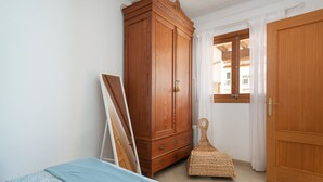 Une chambre à coucher avec un lit, une armoire en bois, une chaise en osier et une fenêtre avec des rideaux.