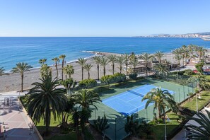 A coastal tennis court surrounded by palm trees, with a beach and ocean in the background.