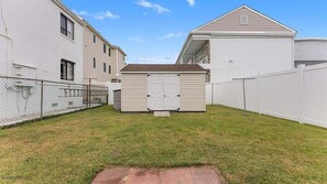 A fenced backyard with a shed, a white fence, and a building with a sloped roof.