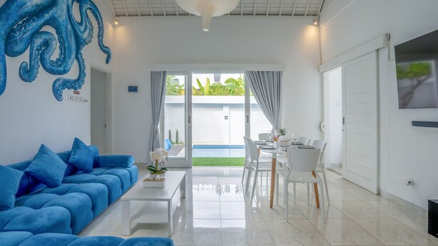 A modern living room with a blue sofa, a dining area with white chairs, and a large window with curtains. - Villa Rio (Canggu)