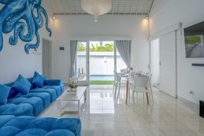 Relax in this spacious living room with a plush sofa, dining area, and large window offering a view of the pool and surrounding greenery.