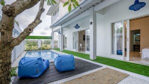 A modern outdoor pool area with a wooden deck, two blue bean bags, and a tree. - Villa Rio (Canggu)