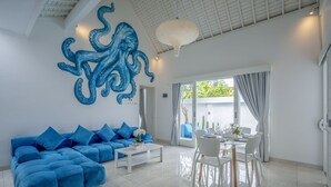 A modern living room with a blue sofa, white coffee table, and a dining area with white chairs. - Villa Rio (Canggu)