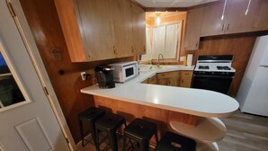 A compact kitchen with wooden cabinets, a white countertop, a microwave, a stove, and three stools. - Woodland cabin on the Bay (Peshtigo)