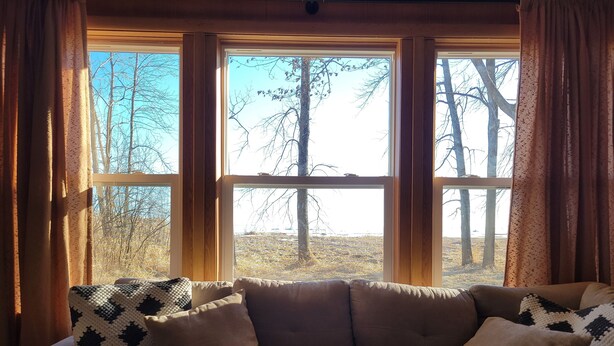 A living room with a beige sofa and patterned throw pillows, large windows with wooden frames, and a view of trees and a field outside. - Woodland cabin on the Bay (Peshtigo)