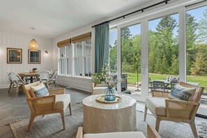 Ett rum med en matplats, flätade stolar, ett runt soffbord och stora fönster med utsikt över en trädgård.