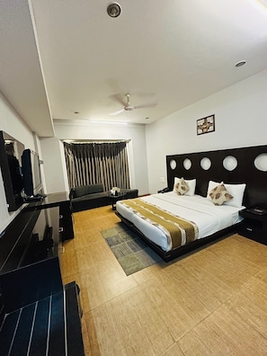 A hotel room with a bed, a desk, a chair, a television, a ceiling fan, and a window with curtains. - Hotel Cymbal-sector 31 Gurgaon (Gurugram)