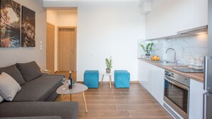 Un salon moderne avec un canapé gris, une petite table ronde et deux poufs bleus. On peut apercevoir une cuisine dotée d’un plan de travail en marbre et d’appareils électroménagers en acier inoxydable.