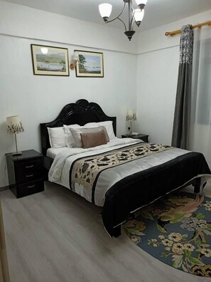 A bedroom with a bed, two nightstands, a lamp, a floral rug, and two framed pictures on the wall. - Beautiful 2-Bed Apartment in Nairobi (Nairobi)