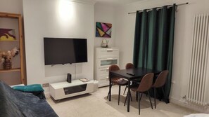A modern living room with a flat-screen TV, a dining table with chairs, a white cabinet, and a green curtain.