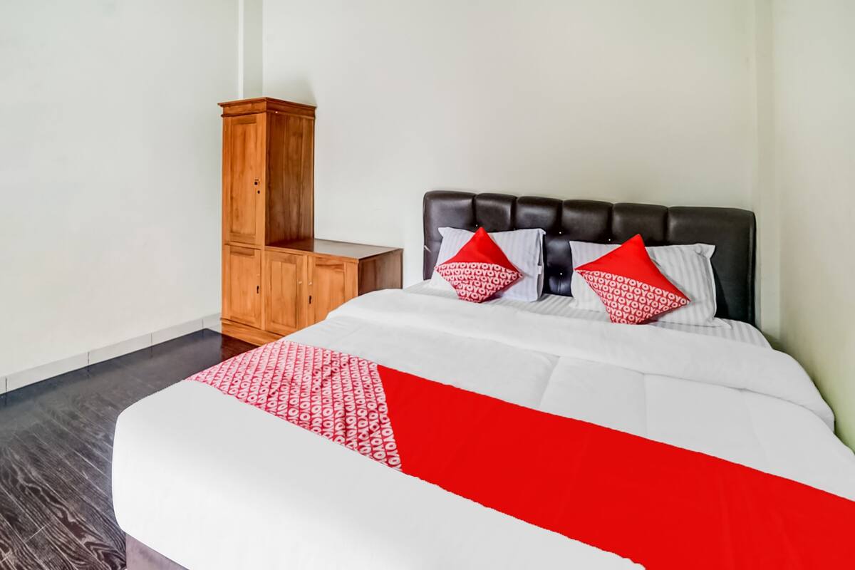 Una cama bien hecha con una colcha blanca y roja, dos almohadas rojas y una mesita de noche de madera.