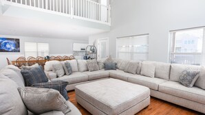 A spacious living room with a large sectional sofa, a wooden coffee table, and a dining area with a white railing. - 5bed/5bath house with heated pool and fire pit! (Murrells Inlet)
