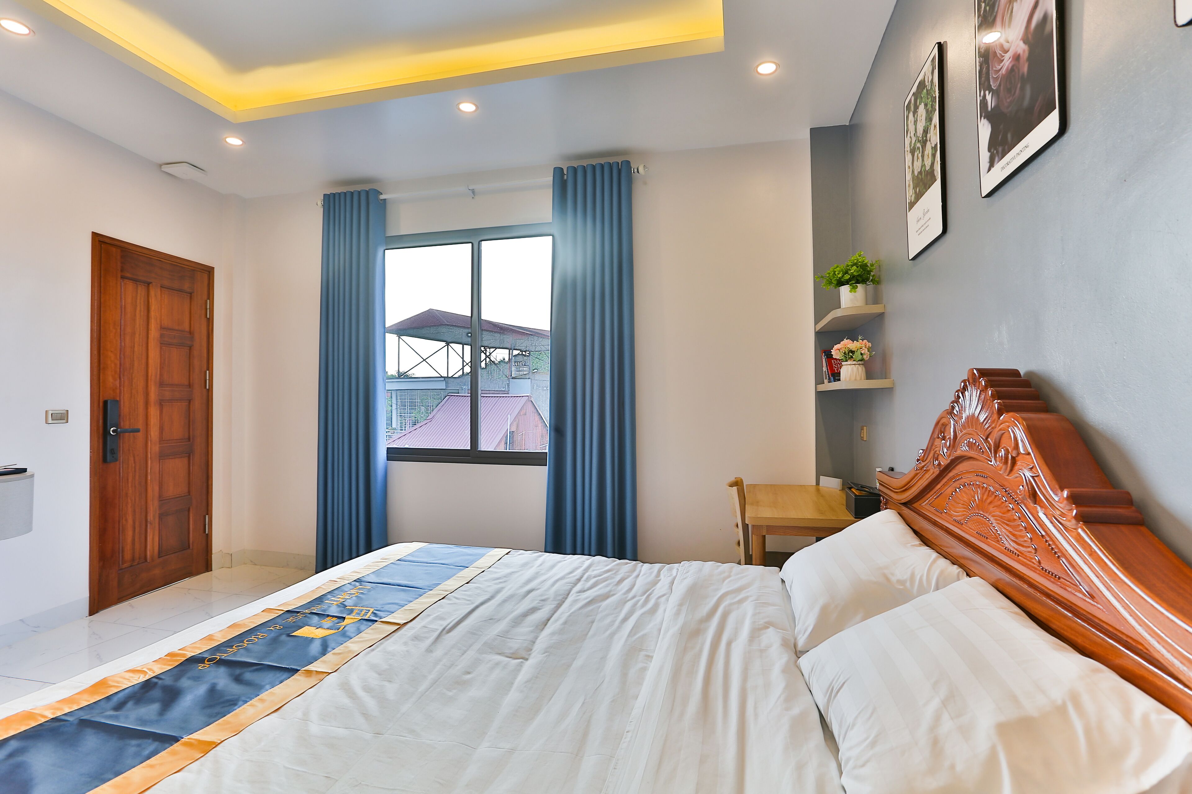 Een slaapkamer met een groot bed, een houten hoofdbord, een raam met gordijnen, een klein tafeltje en een plank met decoratieve objecten.