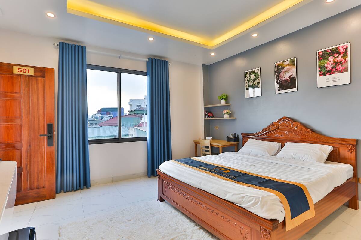 A bedroom with a large bed, a desk, a chair, a wardrobe, and a window with curtains.