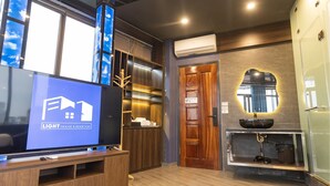 Una habitación de hotel moderna con un televisor grande mostrando un anuncio inmobiliario, muebles de madera, una estantería empotrada y un espejo decorativo.