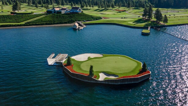 Luftaufnahme eines Golfplatzes mit einem Wasserteil, einem Clubhaus und bewaldeten Hügeln im Hintergrund.