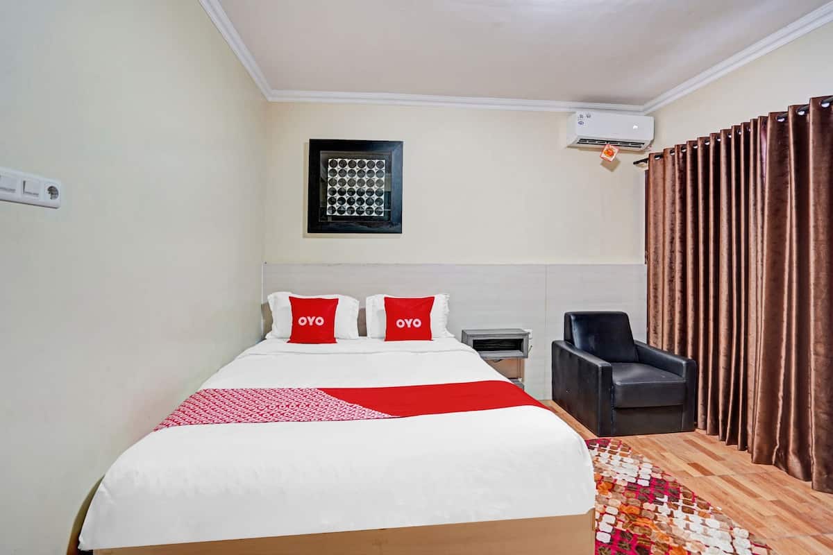 A hotel room with a bed, a chair, a small table, a window with curtains, and an air conditioning unit.