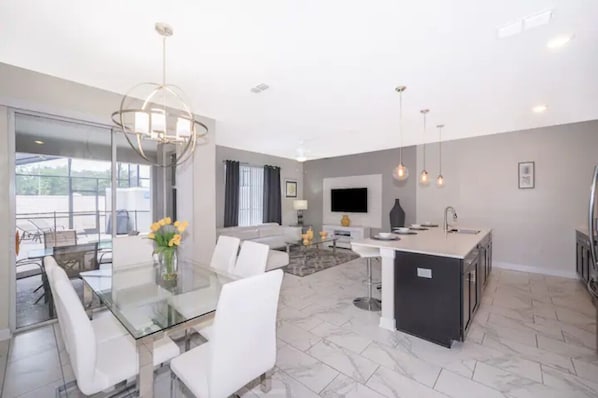 A modern kitchen with a central island, a dining area with a glass table, and a living space with a sofa and TV.