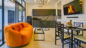A modern living room with a large orange sofa, a glass coffee table, and a dining area with a TV displaying a fire. - Villa Canberra (Seminyak)