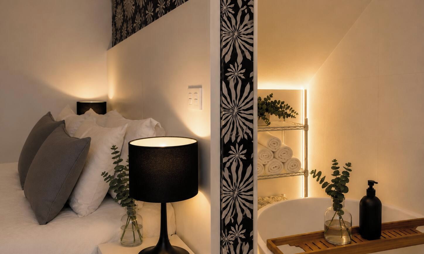 Un dormitorio con cama, una lámpara de noche, una mesita de noche, estante en la pared con toallas y un baño con bañera y bandeja de madera.