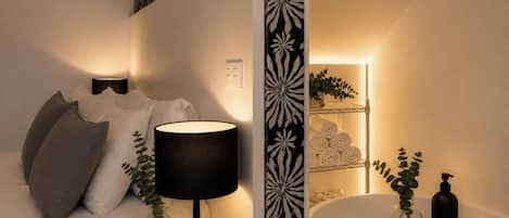 Un dormitorio con cama, una lámpara de noche, una mesita de noche, estante en la pared con toallas y un baño con bañera y bandeja de madera.