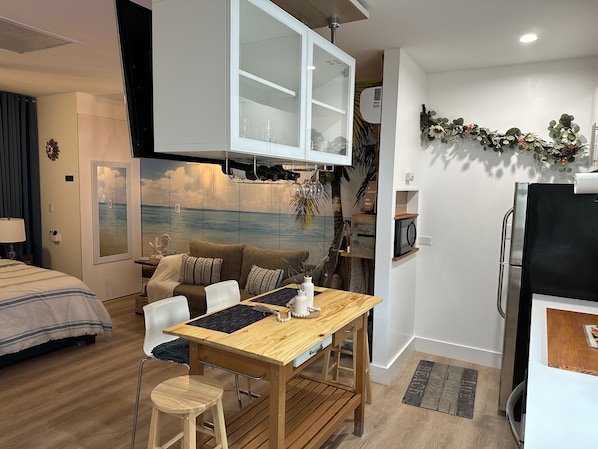 Un espacio habitable compacto con kitchenette, una zona de comedor con mesa y sillas de madera, una cama con ropa de cama a rayas y un televisor montado en la pared.