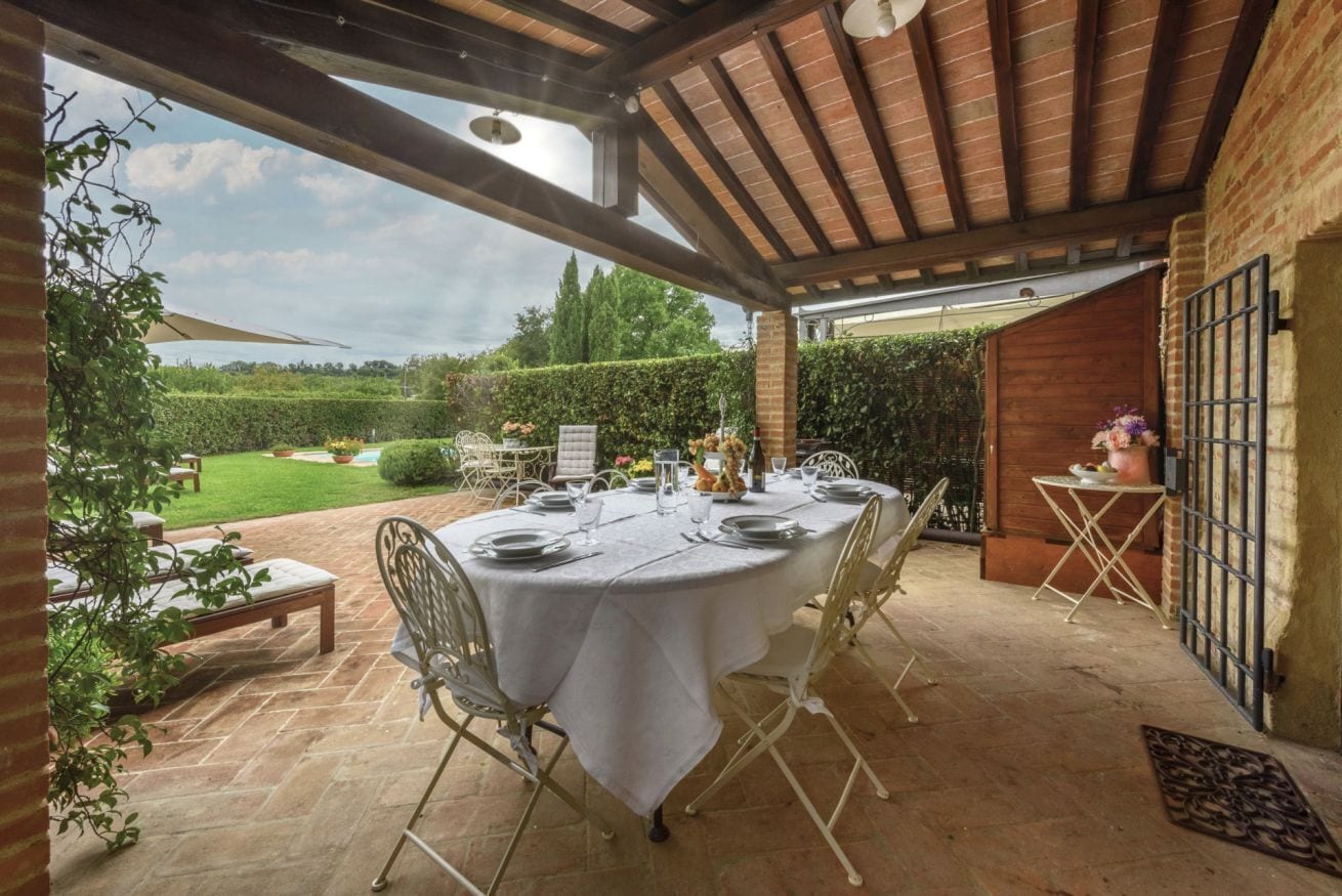 Patio esterno con tavolo rotondo apparecchiato per mangiare, circondato da sedie e una zona coperta con tetto in legno.