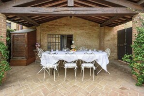 Un espace extérieur abrité, aménagé pour les repas, avec un mur en briques, une porte en bois et une table dressée pour quatre personnes.