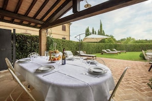 Un espace de restauration en plein air avec une table dressée pour quatre personnes, entourée de chaises, d’une bouteille de vin et d’un panier de fruits.
