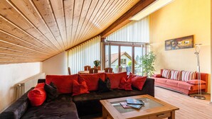 Ein modernes Wohnzimmer mit Holzbalkendecke, einem Ecksofa mit roten Kissen, einem Glastisch und Blick ins Freie.