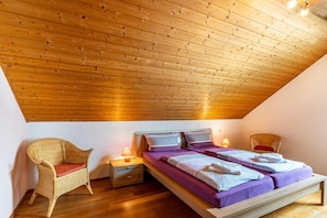 Una habitación con techo inclinado, una cama con ropa de cama morada, una silla de mimbre y una mesita de noche con lámpara.