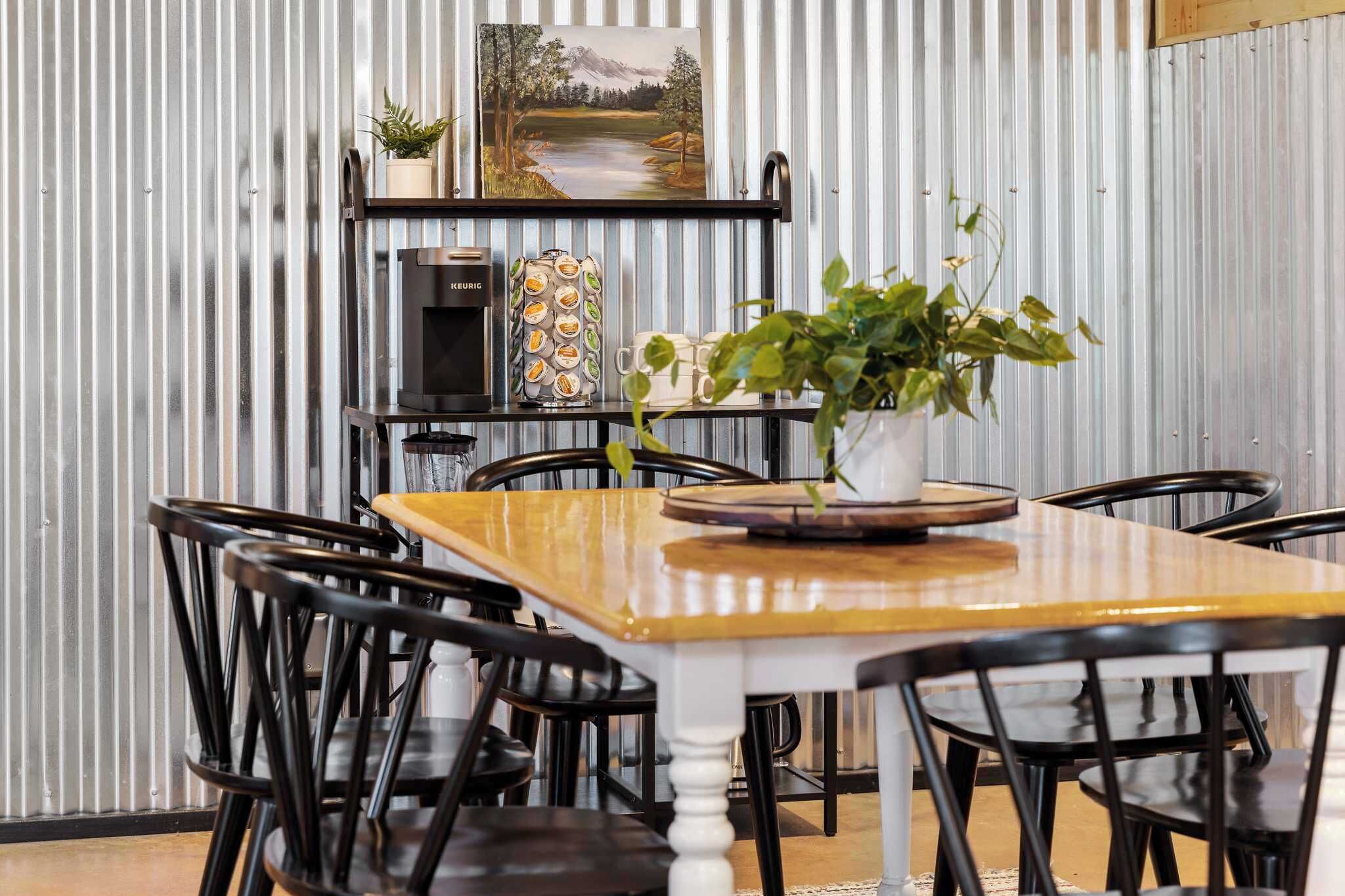 A dining area with a wooden table and black chairs, a coffee machine, a decorative plant, and a shelf with a painting and a coffee display.