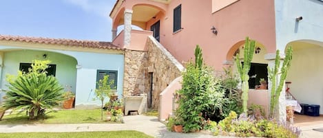 A two-story house with a stone staircase, a small garden with various plants, and a clear blue sky.