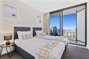 A hotel room with two beds, a nightstand, a lamp, and a balcony with a view of the city and ocean.
