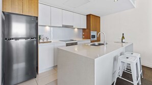 Une cuisine moderne avec un réfrigérateur en acier inoxydable, des armoires blanches, un mur d’accent en bois et un coin bar avec des tabourets.