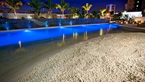 Une piscine avec une plage de galets, entourée de palmiers et de bâtiments modernes.