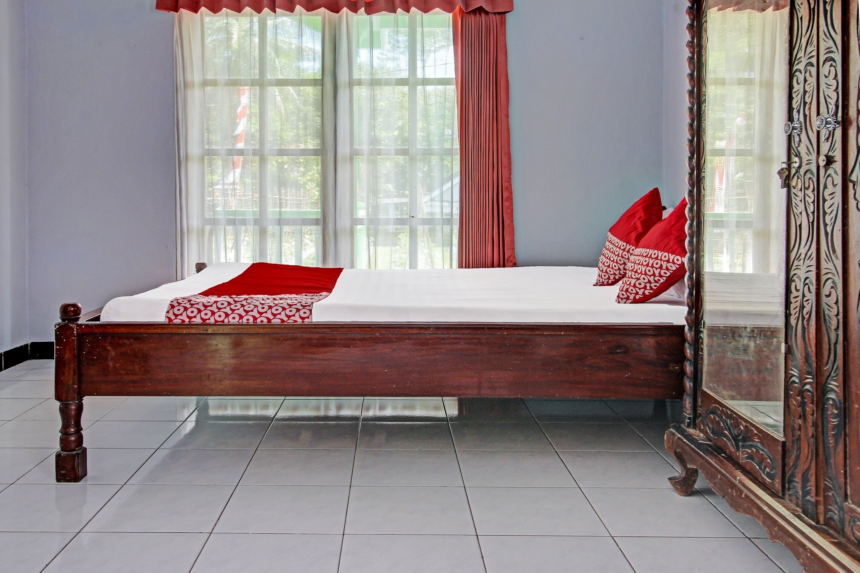 A neatly made bed with red and white bedding, a large mirror with ornate detailing, and a window with sheer curtains.