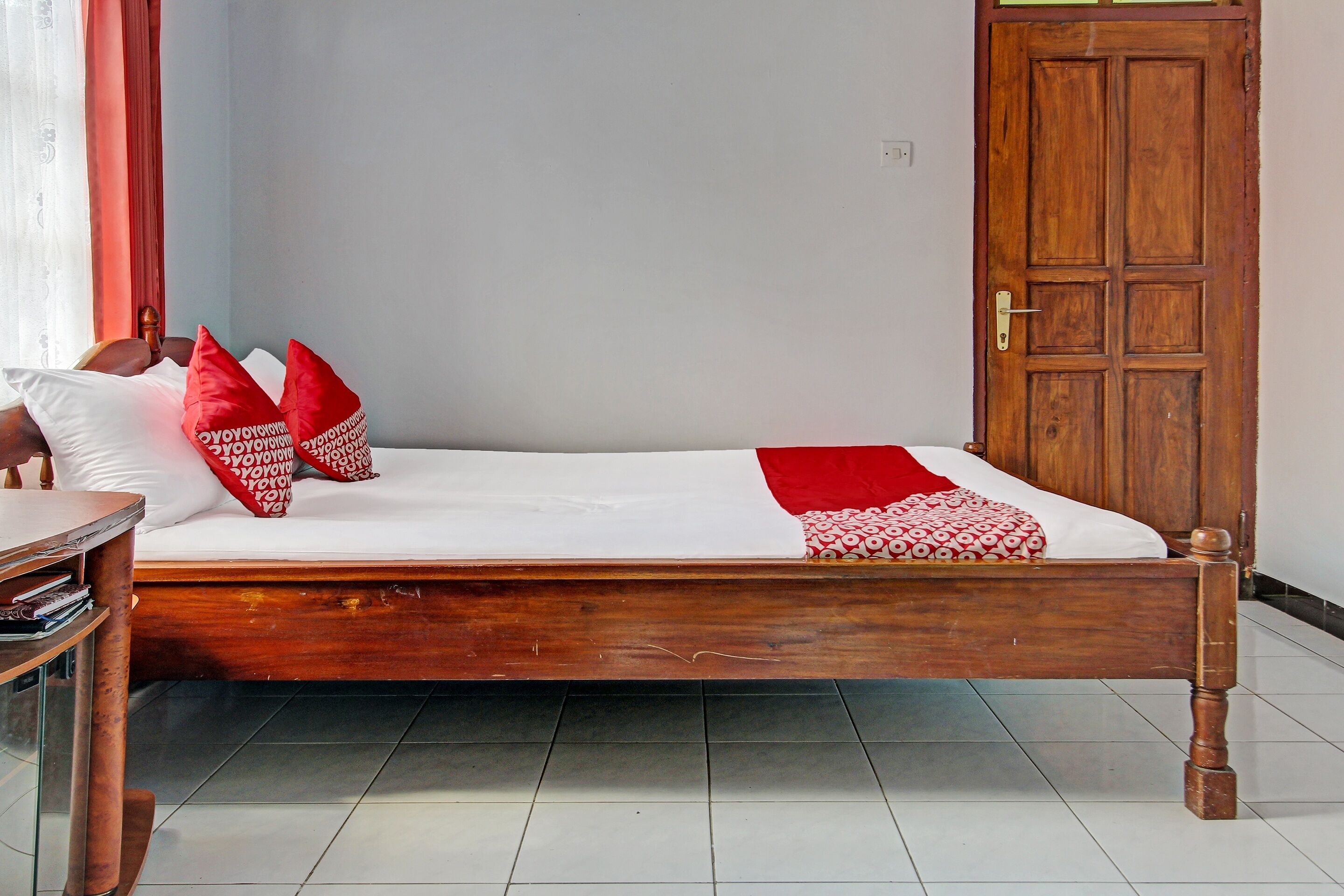 Een eenpersoonsbed met een houten frame, witte beddengoed en rode kussens. Achtergrond toont een houten deur.