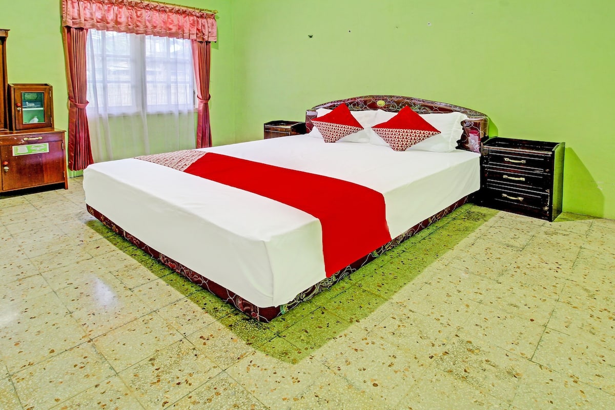 Una cama bien hecha con una colcha roja y blanca, una mesita de noche de madera con cajones, y una pared verde.