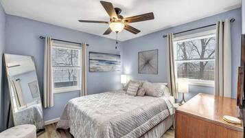 A bedroom with a bed, a ceiling fan, a dresser, and two windows with curtains.