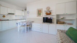 Une cuisine moderne avec des meubles blancs, une table à manger avec des chaises, un téléviseur et des paniers décoratifs placés sur des étagères.