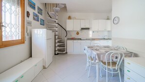Une cuisine de taille réduite avec des meubles blancs, une table à manger avec des chaises, un réfrigérateur et une fenêtre avec des stores.