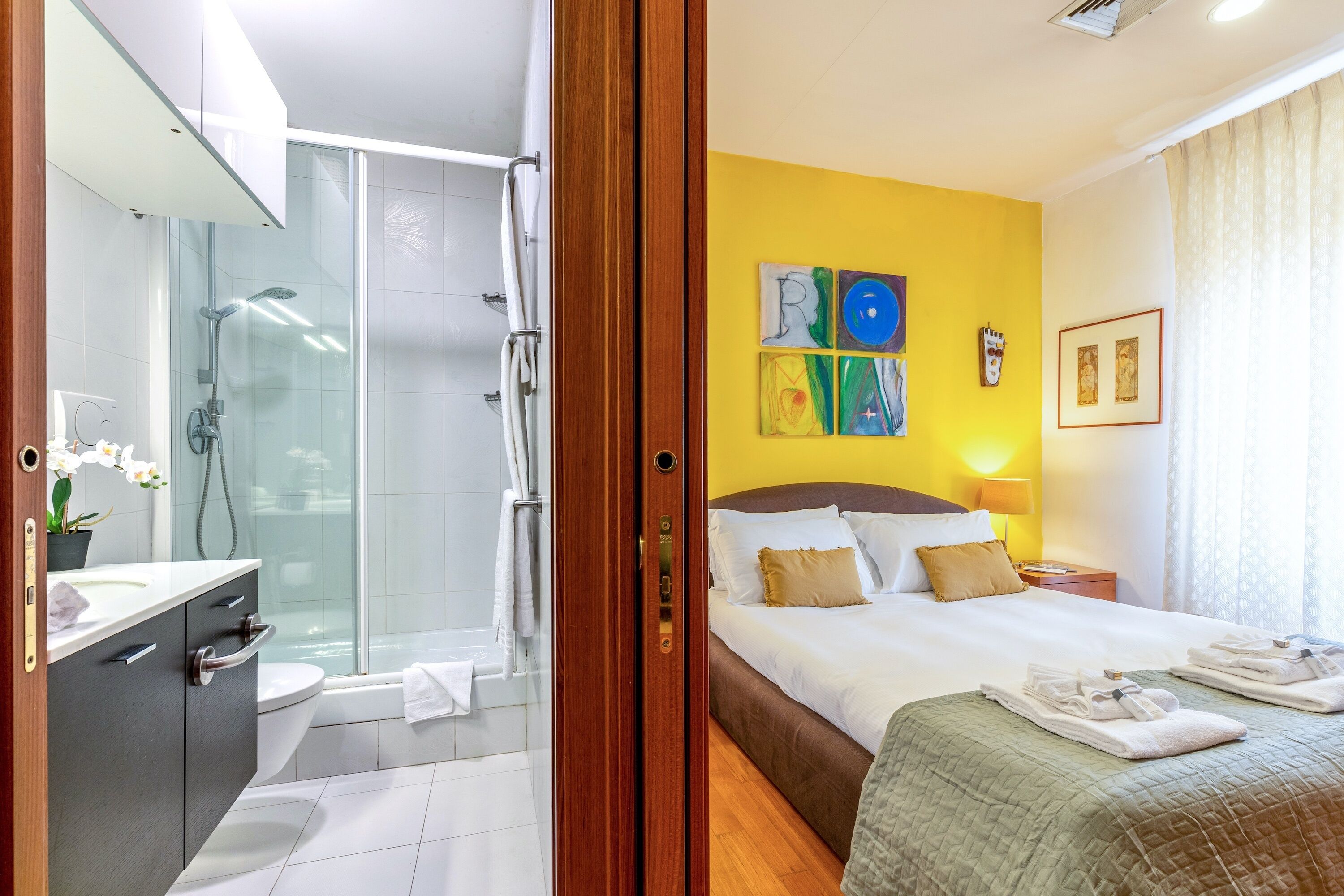 A hotel room with a shower, toilet, sink, bed, and artwork on the wall.