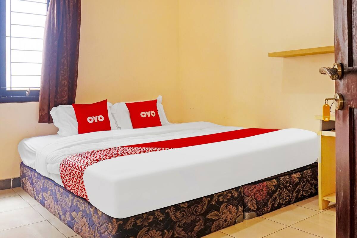 A neatly made bed with a red and white bedspread, two pillows with the "OYO" logo, and a patterned bed skirt.
