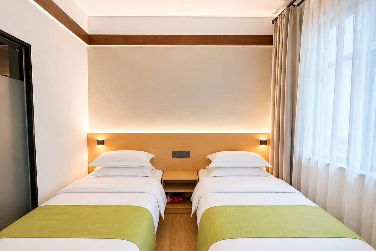 A hotel room with two single beds, wooden headboard, bedside lamps, and a window with curtains.