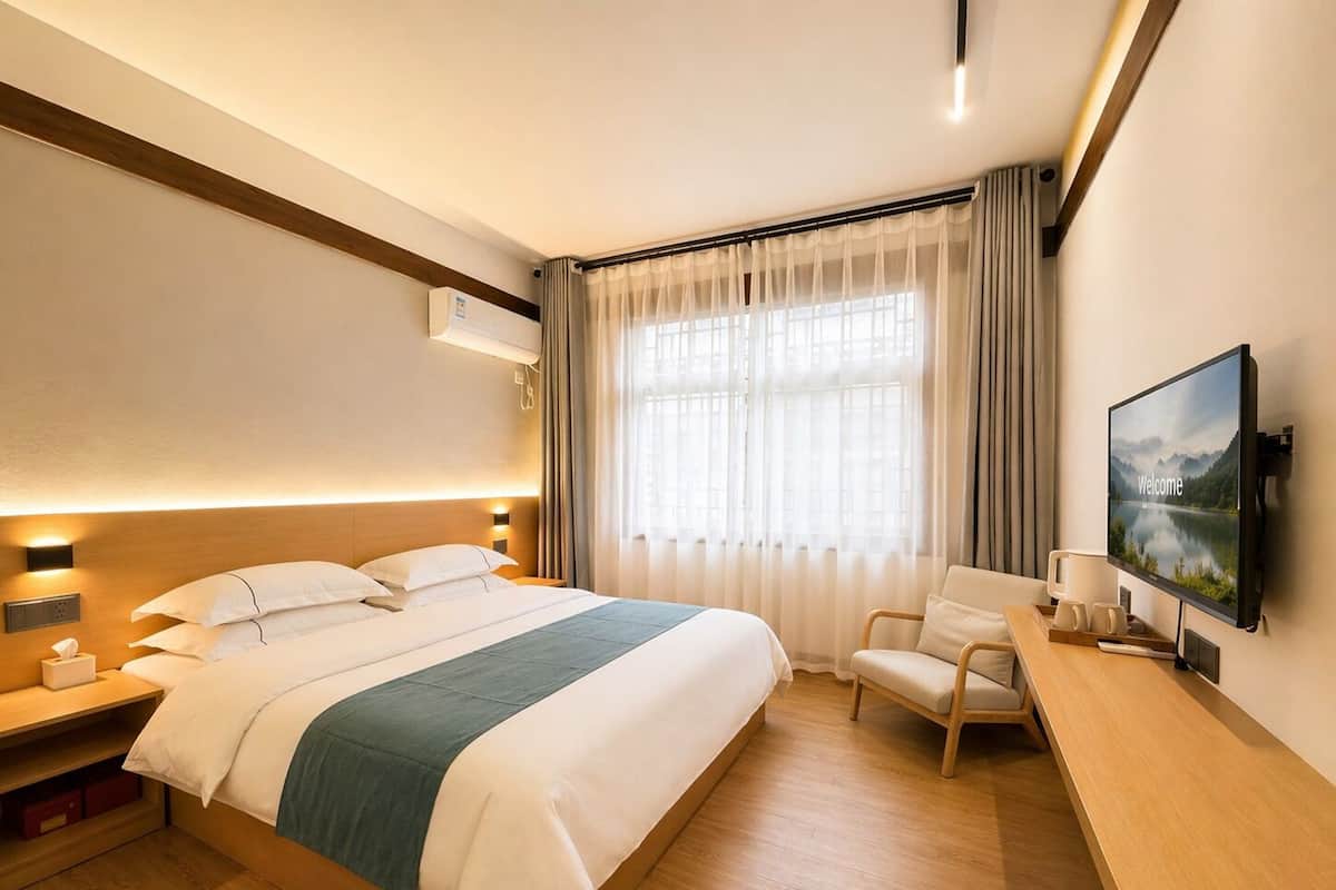 A hotel room with a bed, bedside table, chair, TV, and window with curtains.