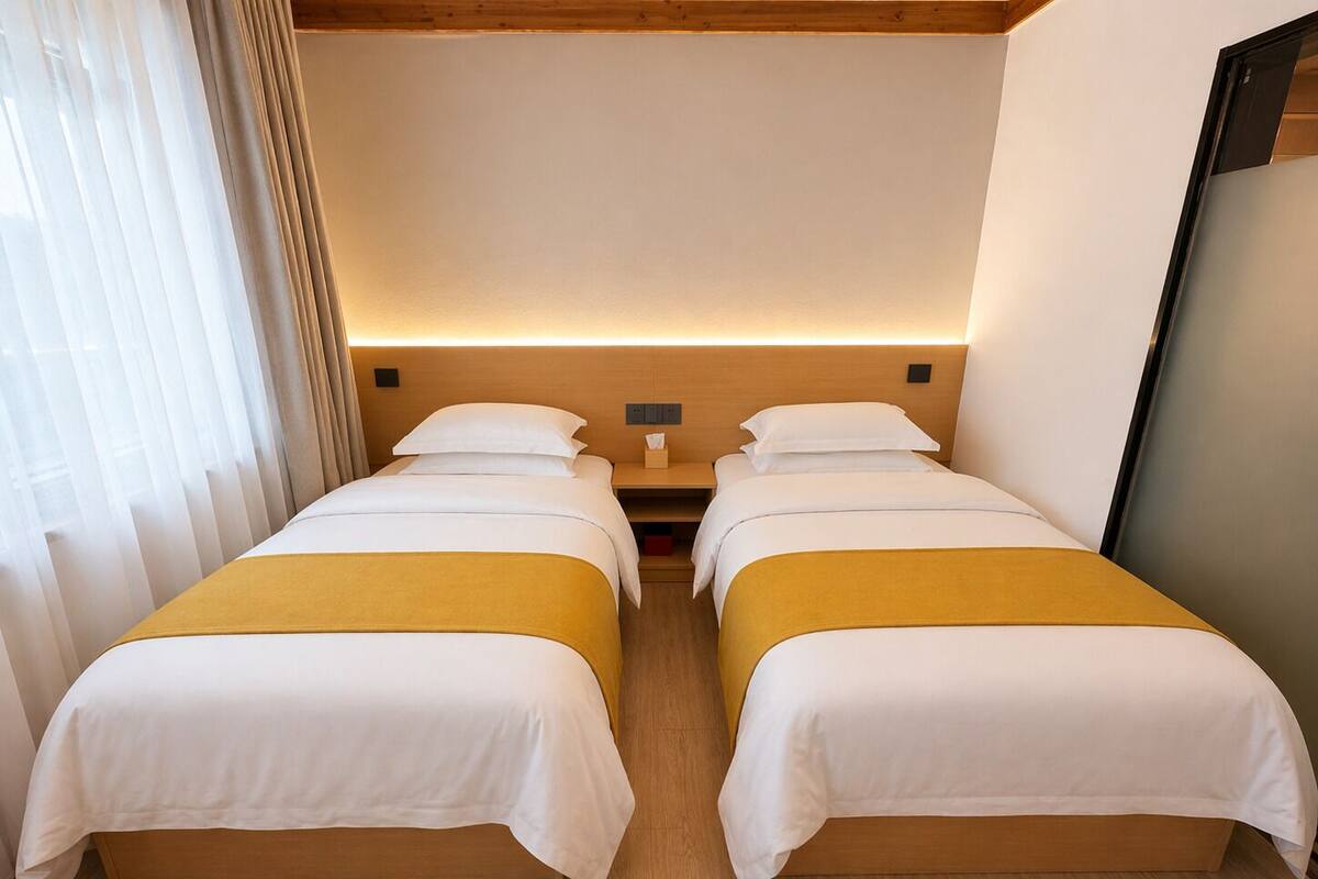A hotel room with two beds, wooden headboard, bedside tables, and a large window with curtains.