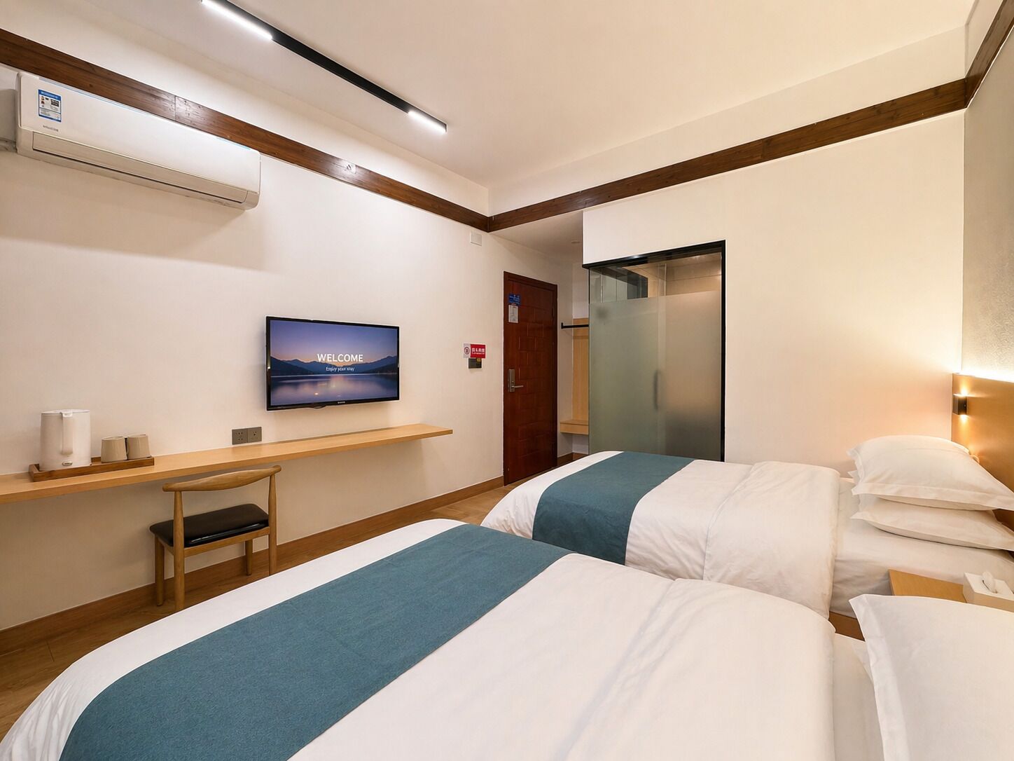 Hotelkamer met twee bedden, een televisie waarop een welkomstbericht staat, een bureau met een stoel en een airconditioning.